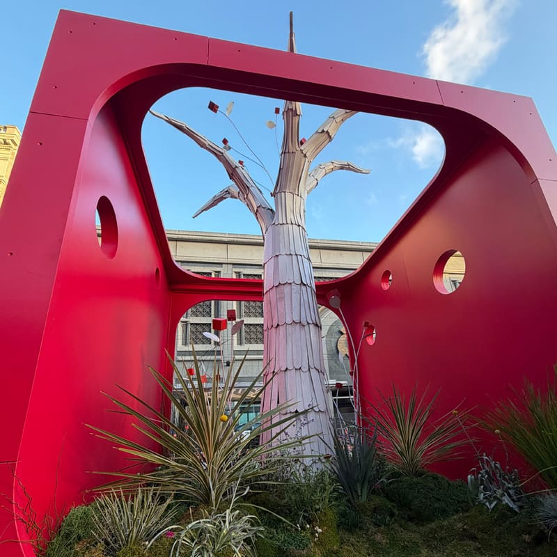 Community Tree by Musepop: a white tree rising through a red cube against a blue sky, with grasses and ferns at its base.